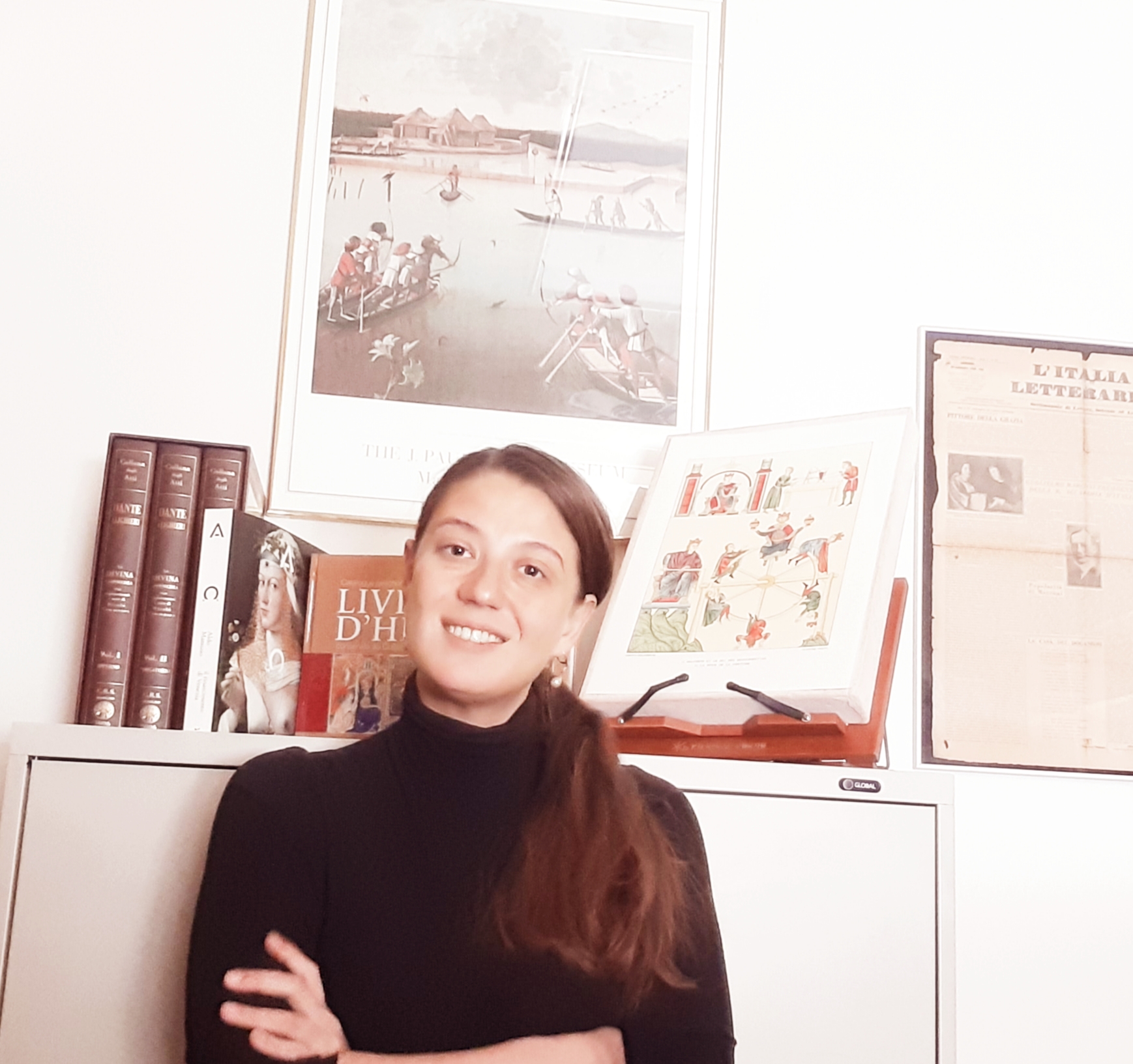 colour headshot photo of Elisa Brilli against beige filing cabinet with books and illustrated wall hangings.