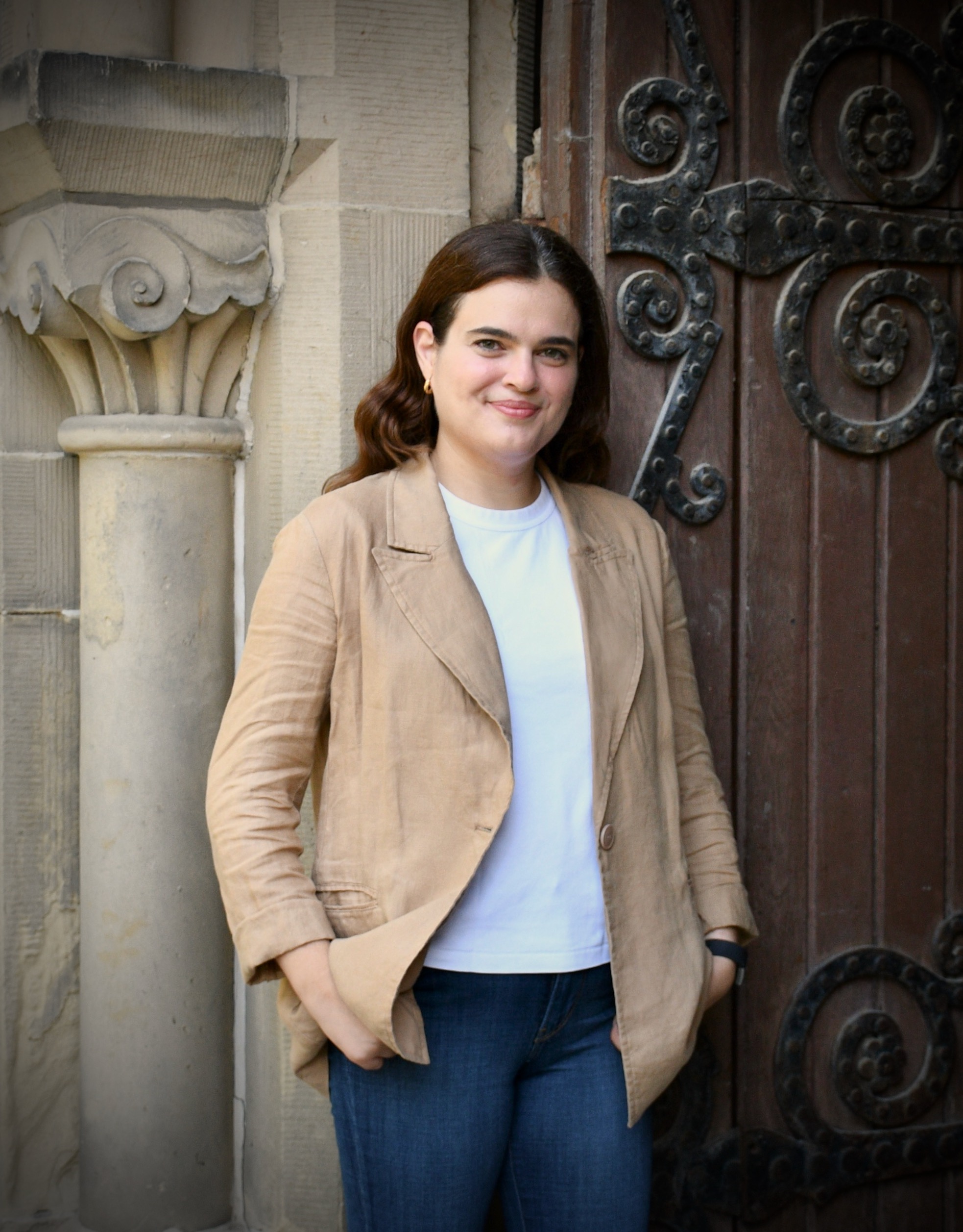 Colour 3/4 bodyshot of Laura Ingallinella standing outside against a stone carved column and wooden door with wrought iron plating