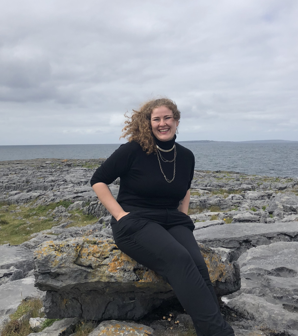 Colour photo of a smiling Olivia Cox with hands in pants pockets in black turtleneck, pants, three strands of silver colour necklaces, with windswept curly brown hair, seated on a boulder outdoors on a rocky shoreline.