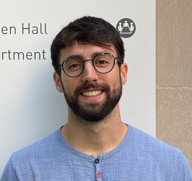 Colour headshot photo of Mattia Ragazzoni wearing eye glasses, blue shirt, short brown hair, outdoors against a billboard.