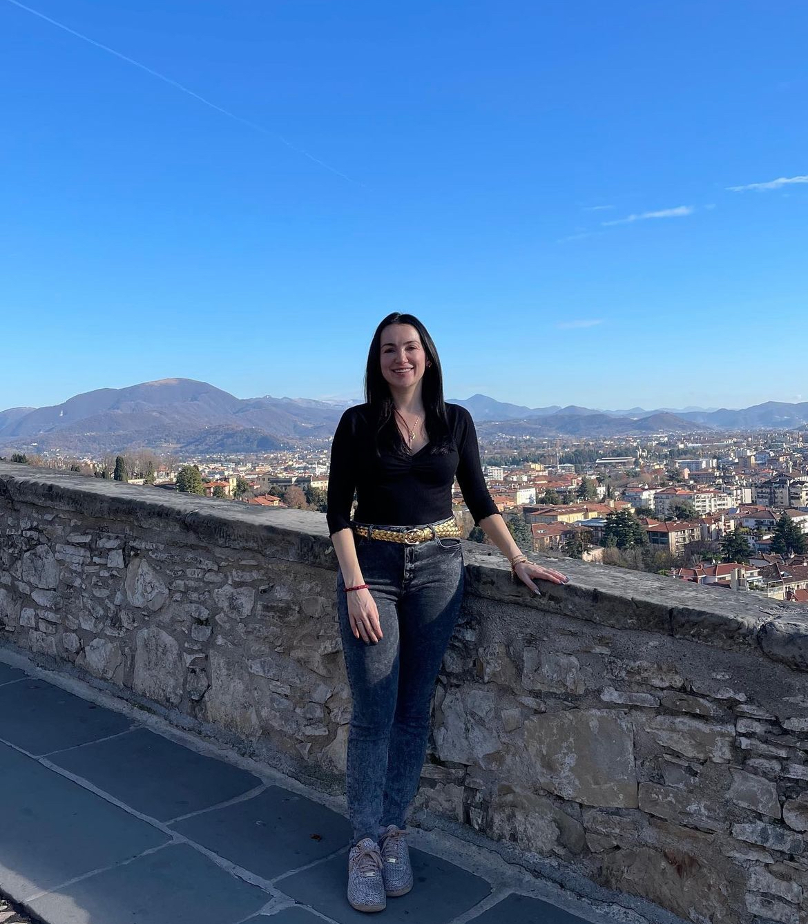 Full length portrait of Rachel Grasso outdoors against waist height stone wall with background of low-rise buildings in Bergamo, Italy. 