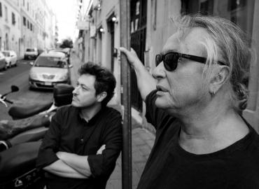 black and white photo of Porpora Marcasciano wearing sunglasses right hand on street pole and Vittorio Martone leaning on same street pole, both in side profile on a street in Rome during the shooting of the documentary “Porpora&amp;quot;