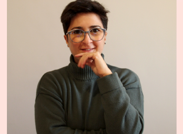 picture of Anna Mongibello, wearing a dark sweater, grey glasses, holding her chin, short hair, smiling. Against a grey and pink background.