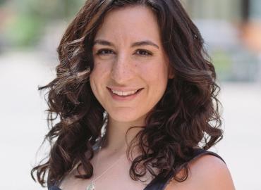 An image of a 30 year old white woman with dark shoulder length curly hair. Her face and upper torso is pictured. She is smiling and looking into the camera. She is wearing a blue denim dress with her arms exposed. She is standing outside on a sunny day, 