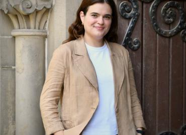 Colour 3/4 bodyshot of Laura Ingallinella standing outside against a stone carved column and wooden door with wrought iron plating